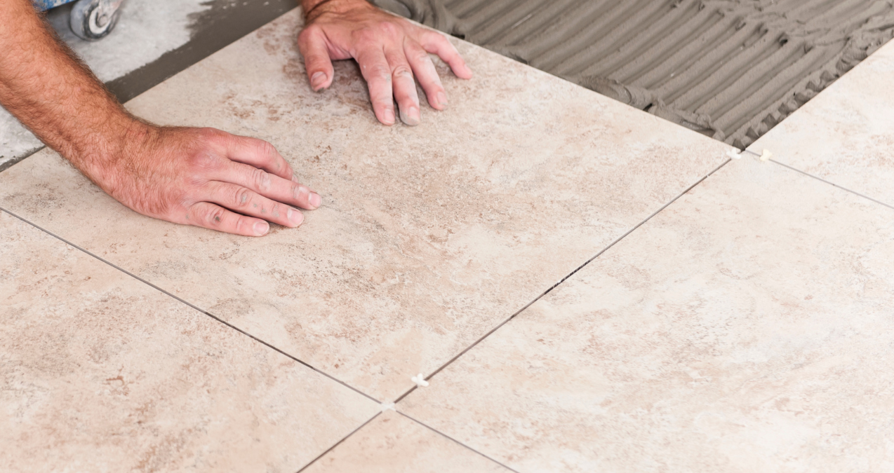 Person placing beige floor tile, hands on surface with gray adhesive.