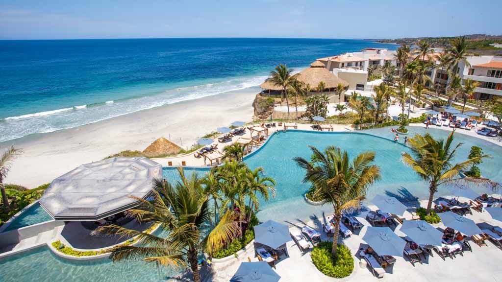 An aerial view of a beach resort with a large swimming pool surrounded by palm trees.