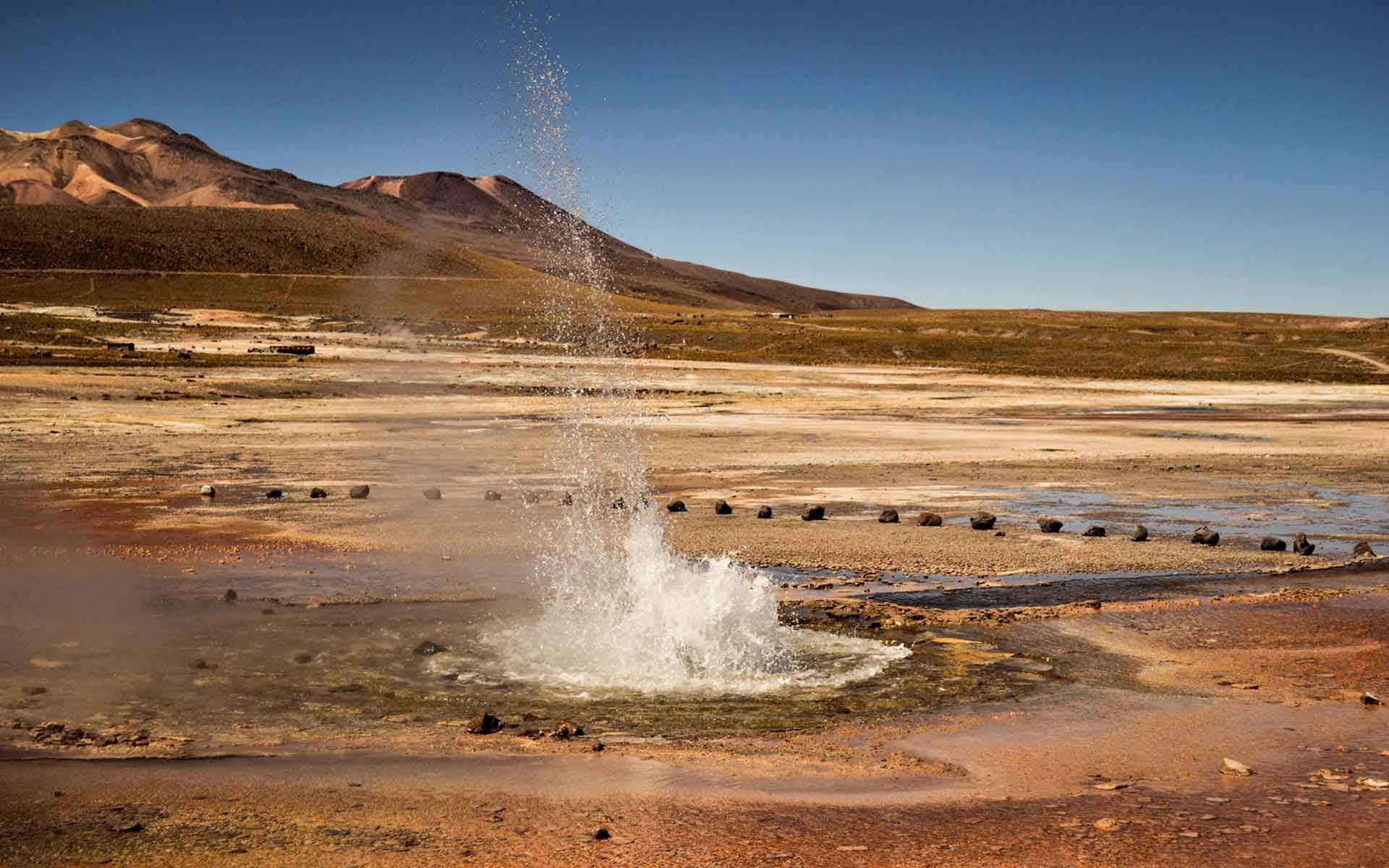 A geyser is erupting in the desert with mountains in the background.