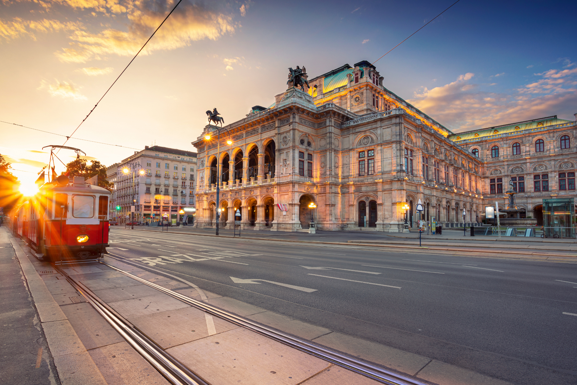 A red trolley is driving down a street in front of a large building at sunset.