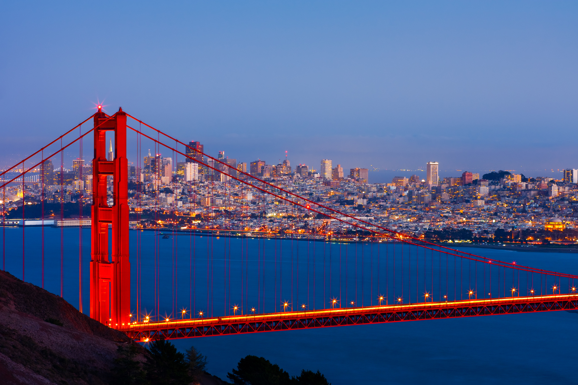 The golden gate bridge is lit up at night with a city in the background.