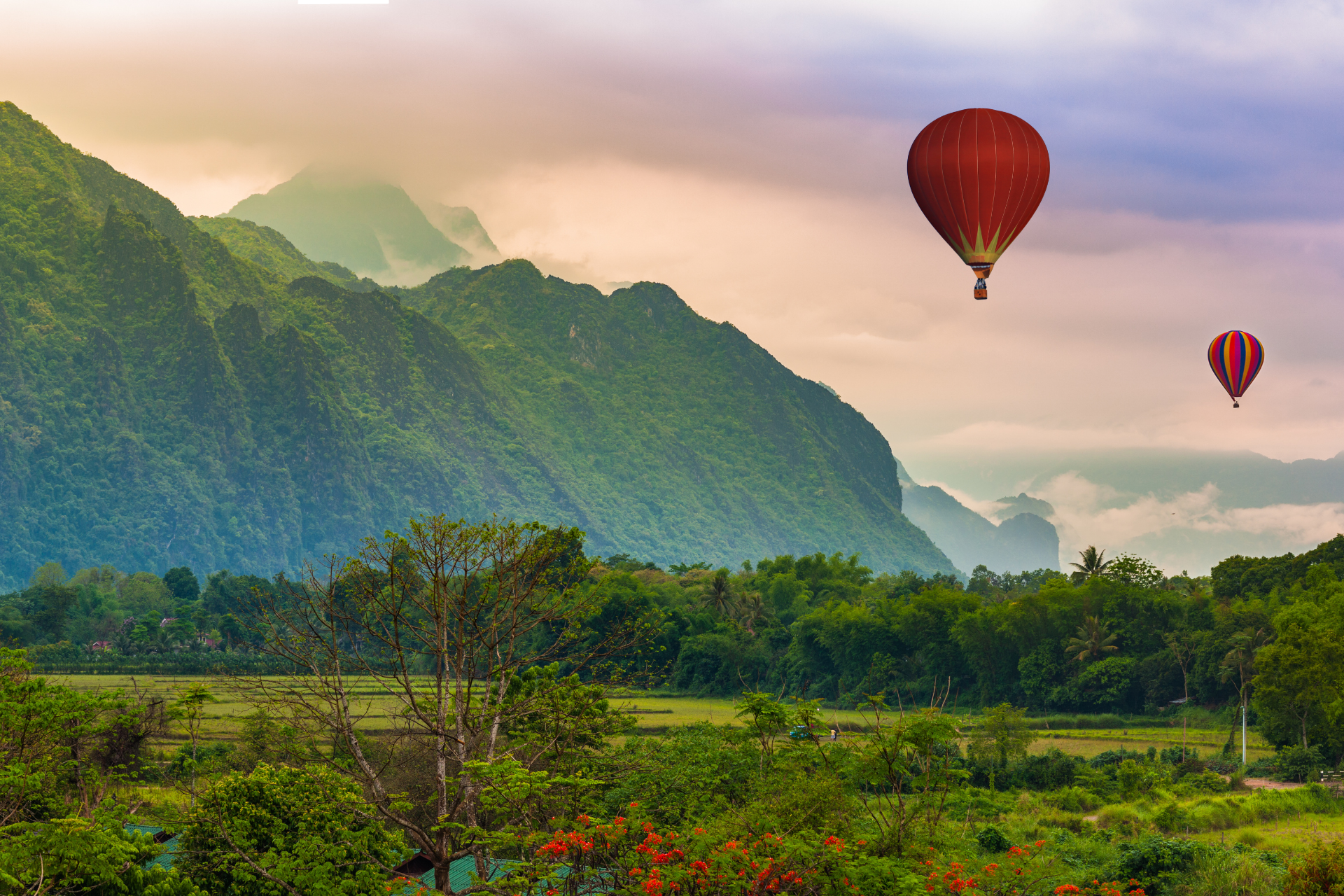 Two hot air balloons are flying over a field and mountains.