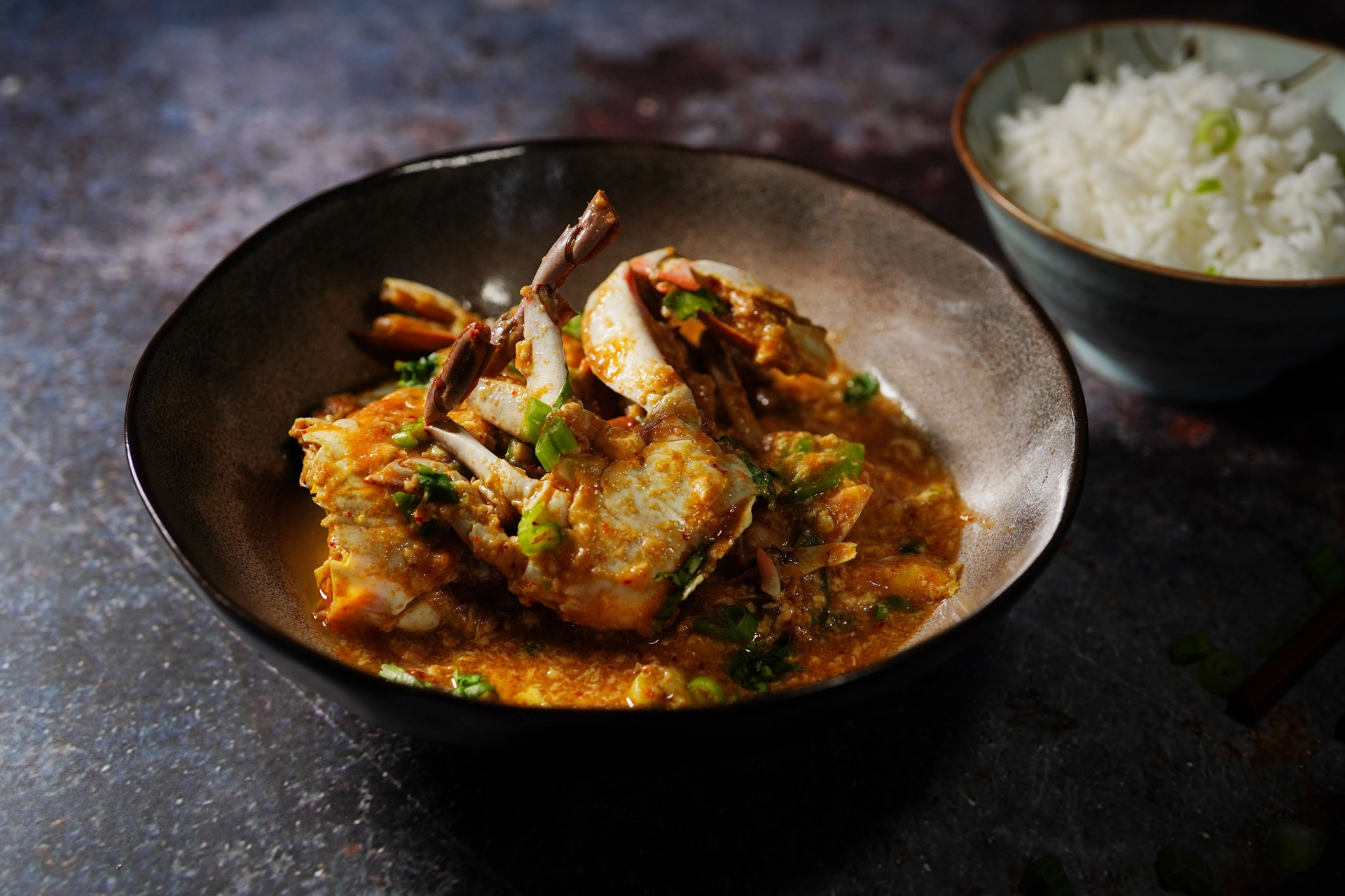 A bowl of crab curry next to a bowl of rice on a table.