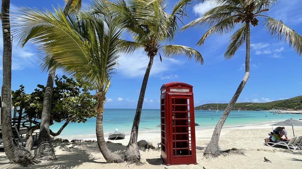 A red telephone booth is sitting on a beach surrounded by palm trees.