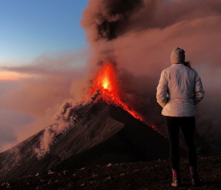 A woman in a white jacket stands in front of a volcano