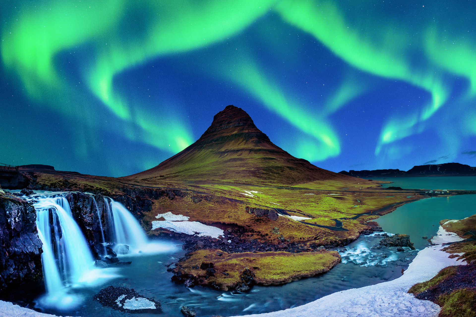 The aurora borealis is shining over a waterfall and a mountain.