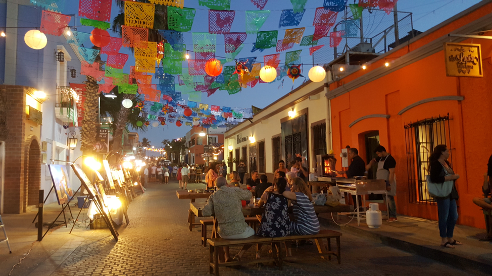 A group of people are sitting at tables in a narrow street with colorful flags hanging from the ceiling.