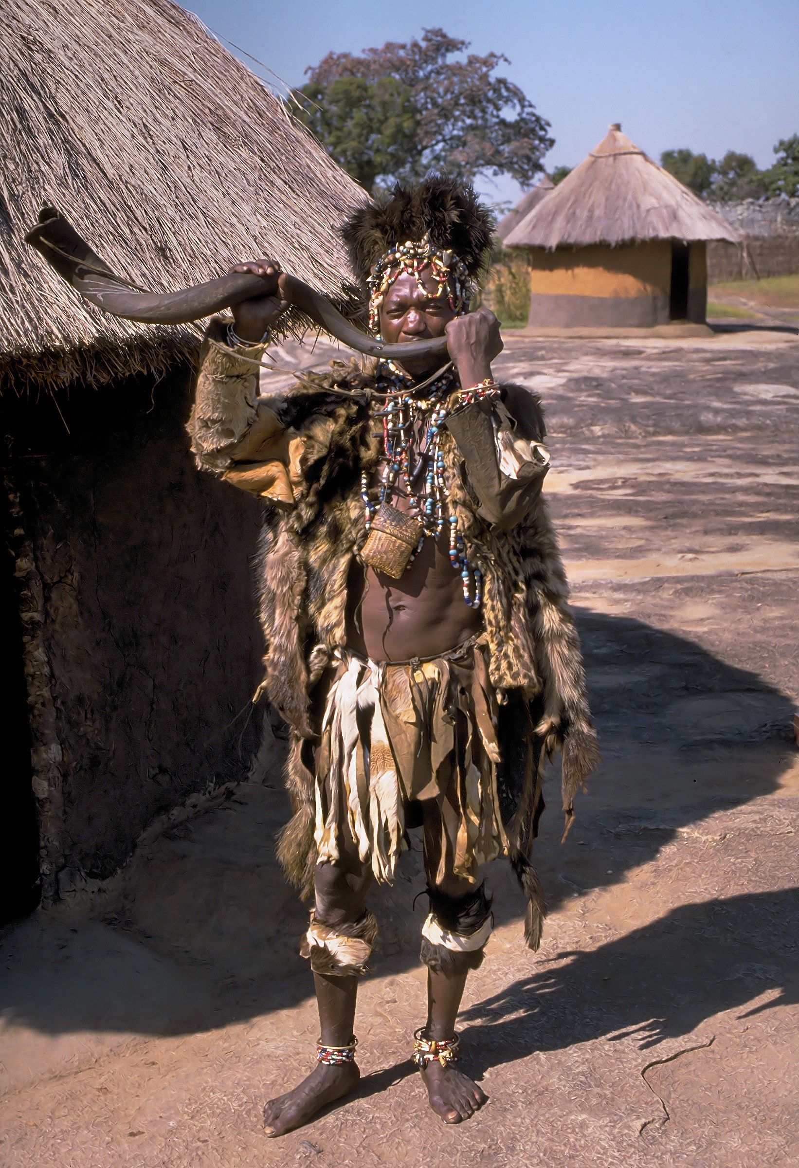 A man in a traditional costume is holding a horn in front of a thatched hut.