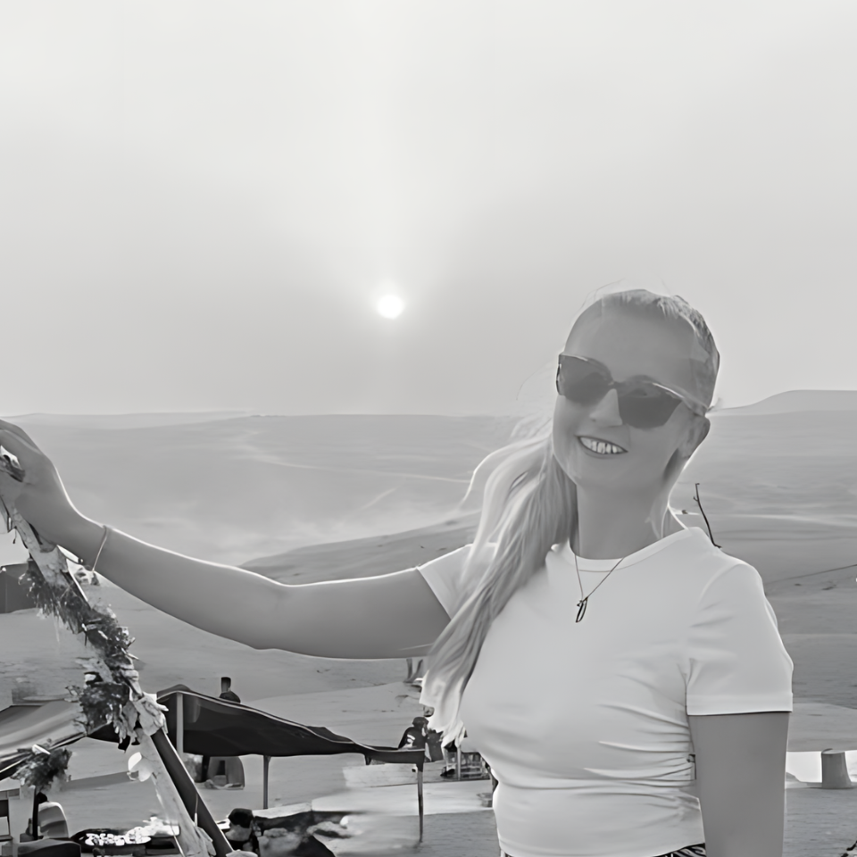 A woman wearing sunglasses and a white shirt is standing in front of the ocean.