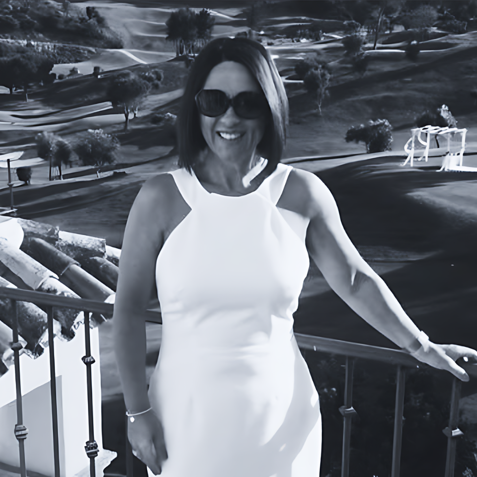 A woman in a white dress and sunglasses stands on a balcony overlooking a golf course