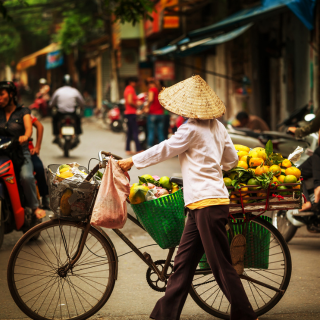 A woman wearing a conical hat is pushing a bicycle full of fruit