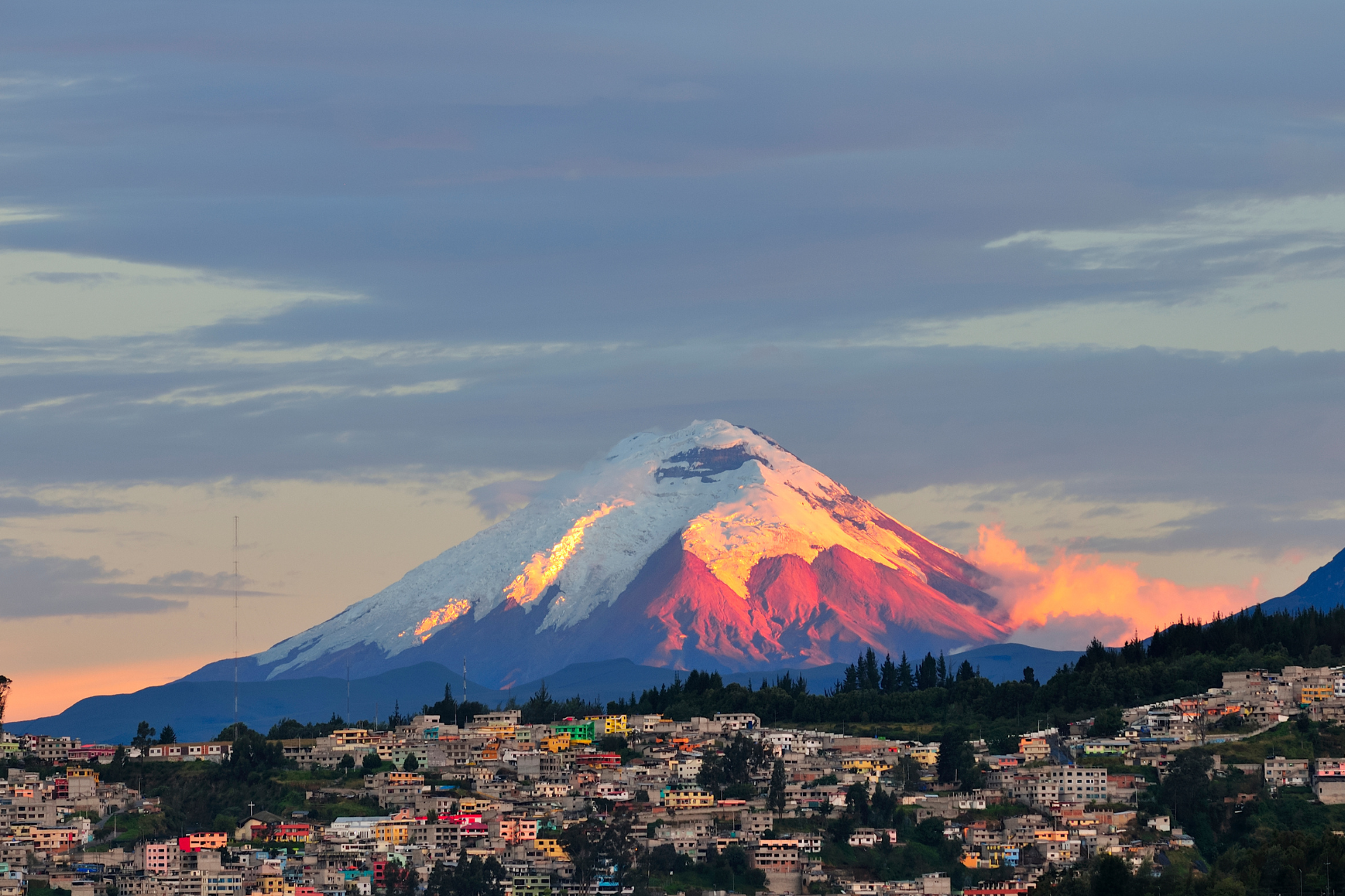 A mountain covered in snow is surrounded by a city at sunset.