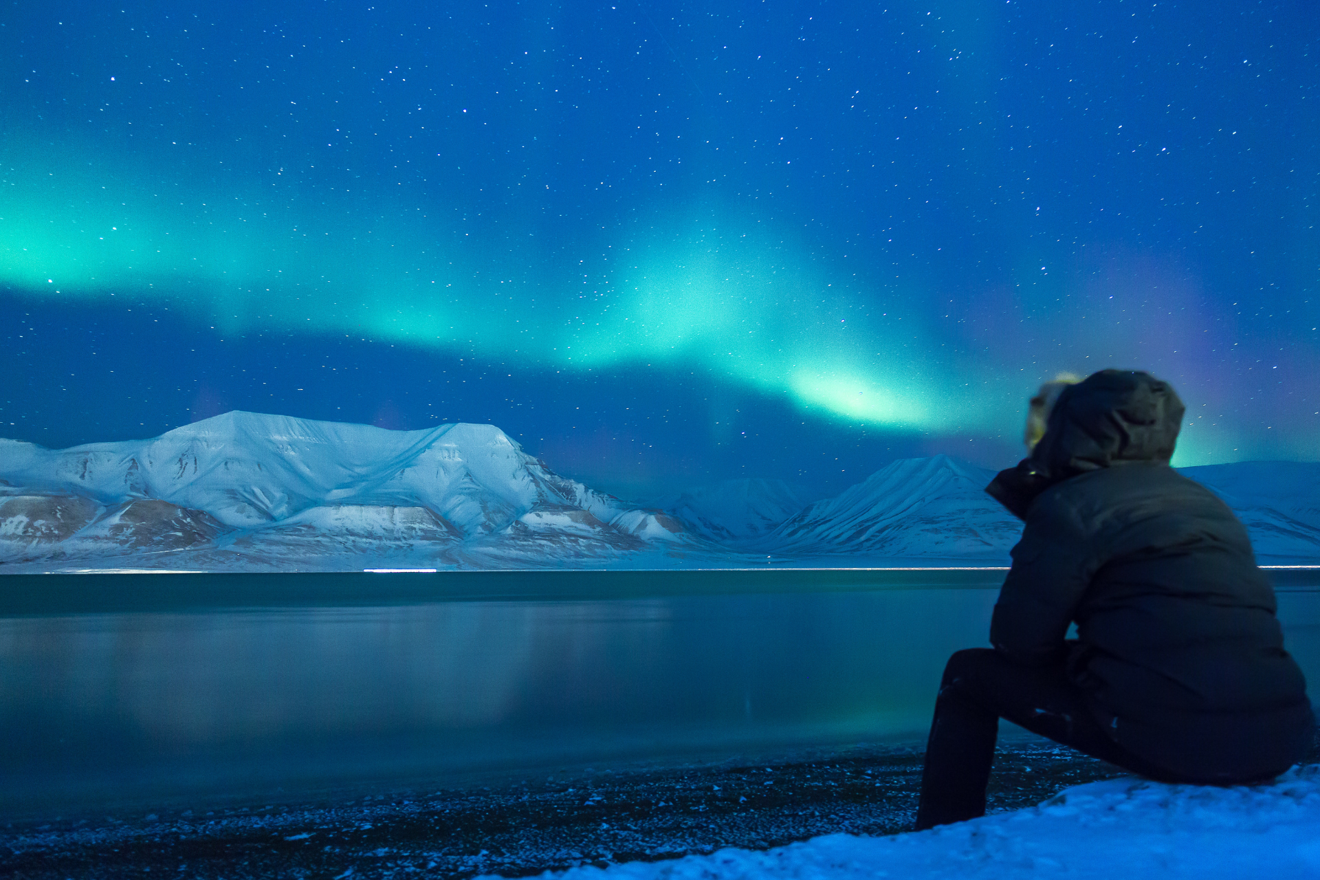 A person is sitting on the shore of a lake looking at the aurora borealis.