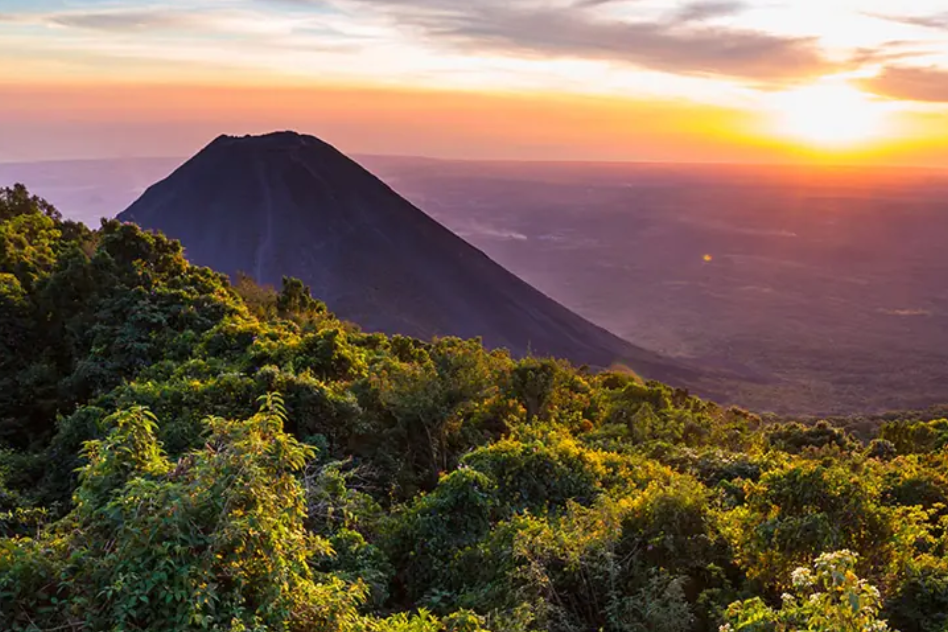 A volcano with a sunset in the background and a lush green forest in the foreground.