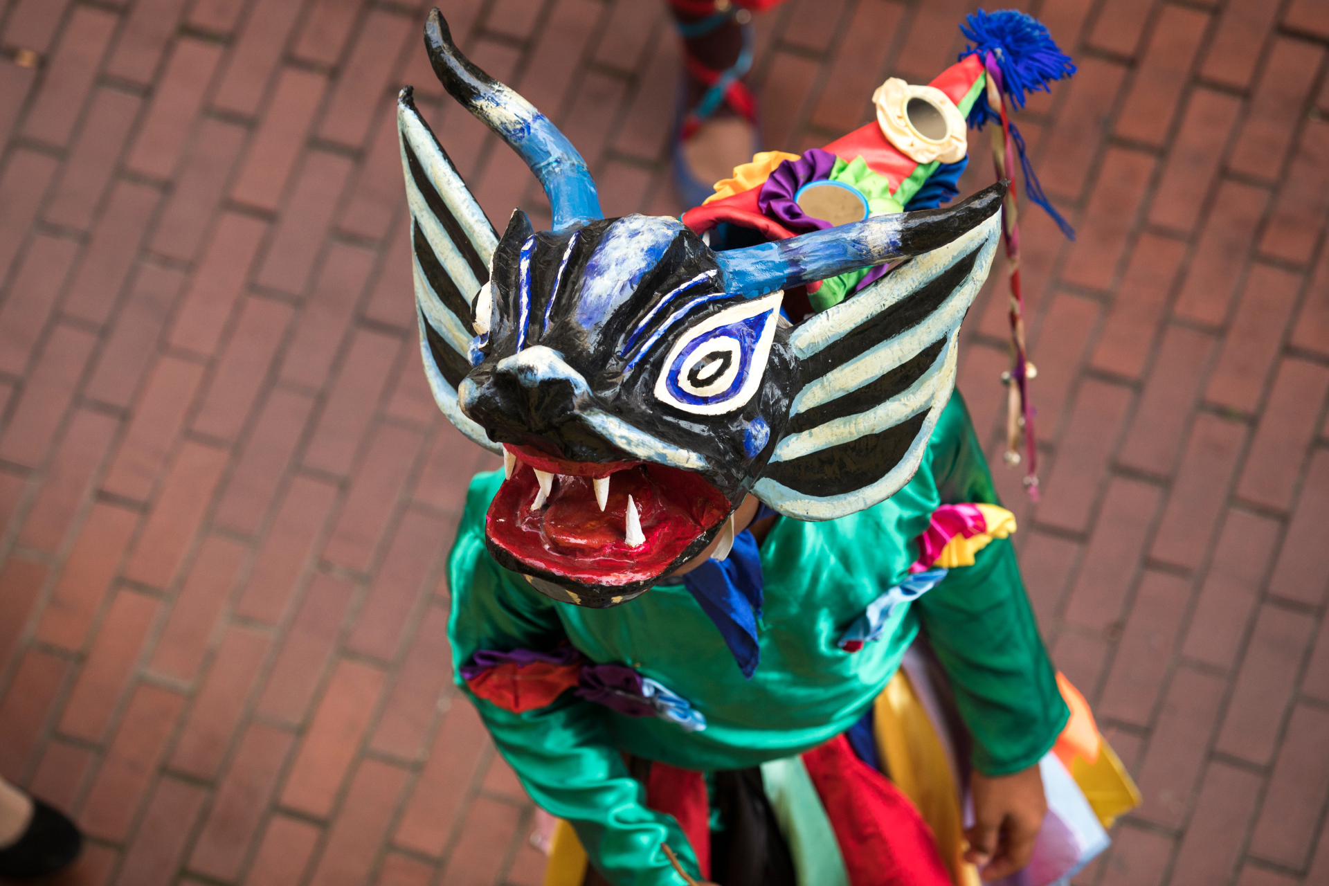 A person wearing a colorful dragon mask is standing on a brick sidewalk.