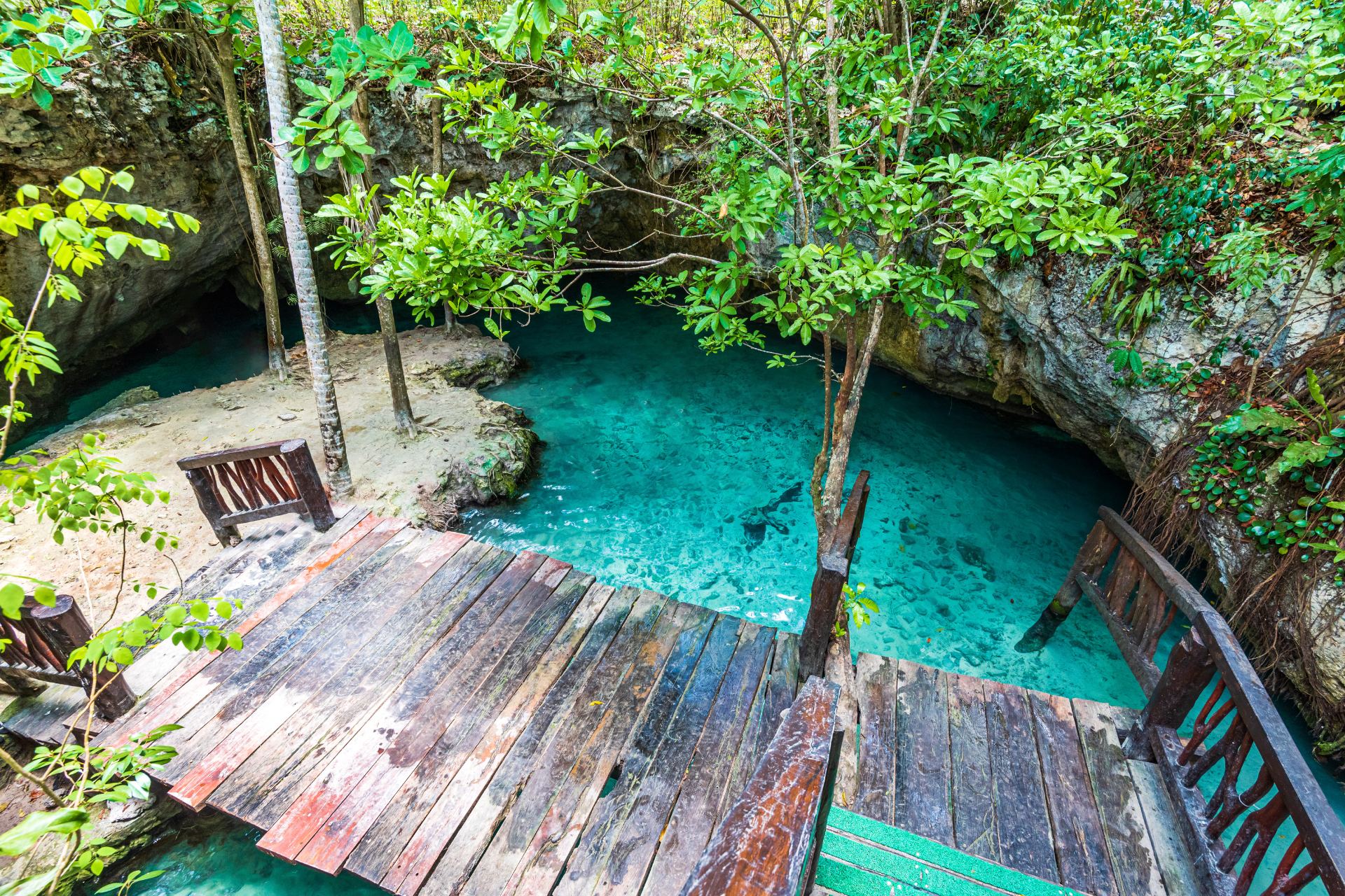 A wooden bridge leading to a blue pool in the middle of a forest.