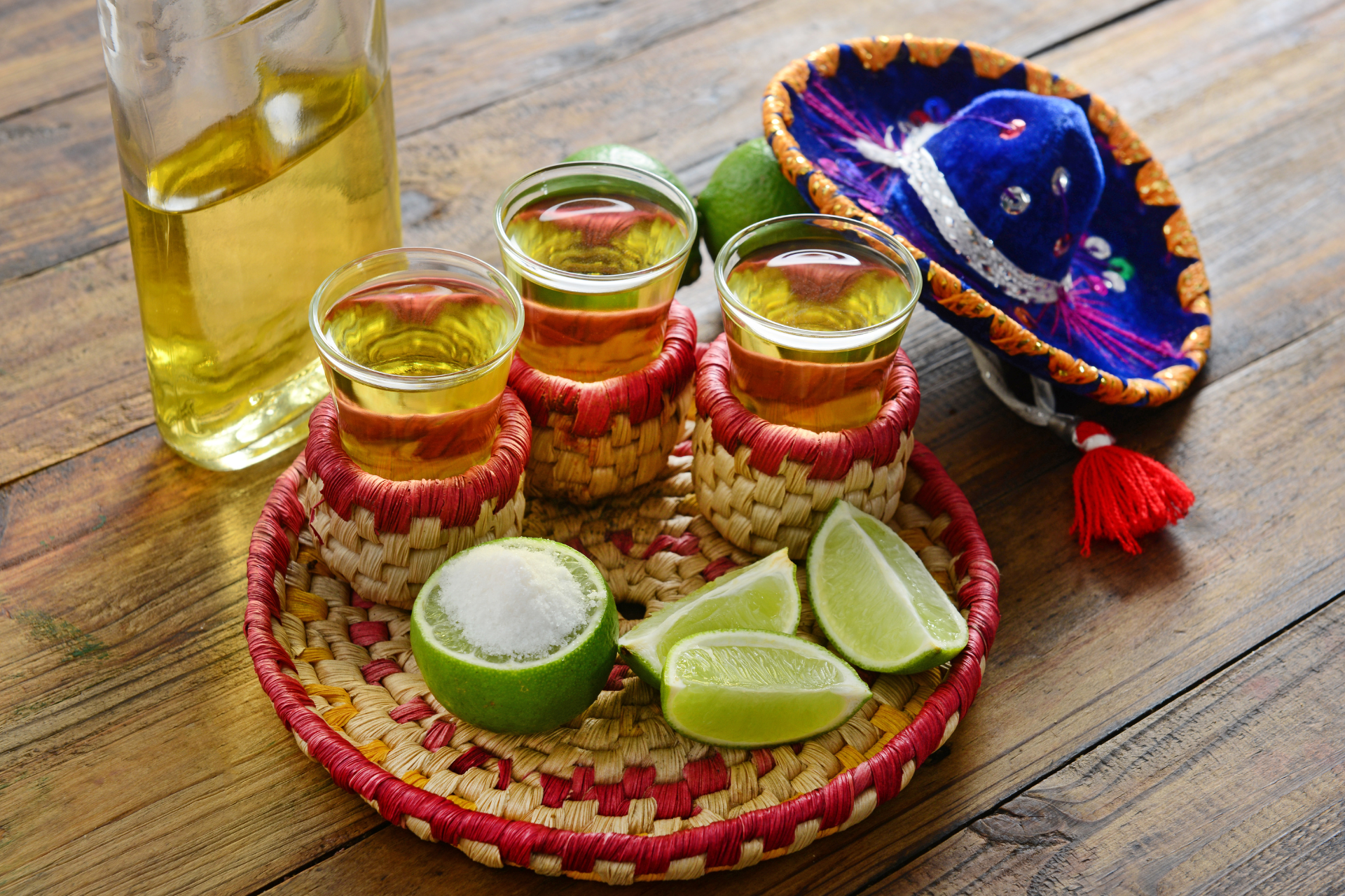 A tray of tequila , limes , salt and a sombrero on a wooden table.