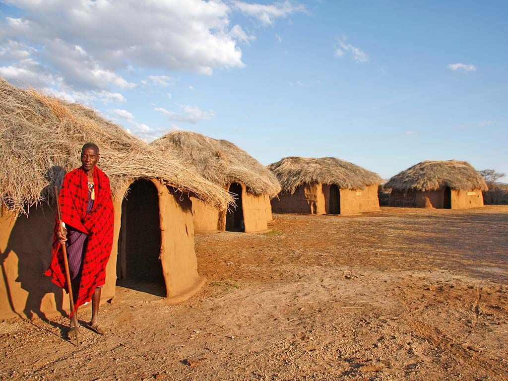 A man in a red robe is standing in front of a thatched hut.