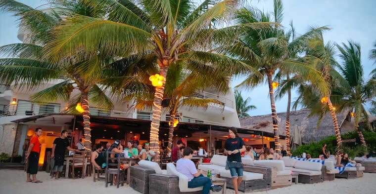 A group of people are sitting at tables on the beach in front of a restaurant.