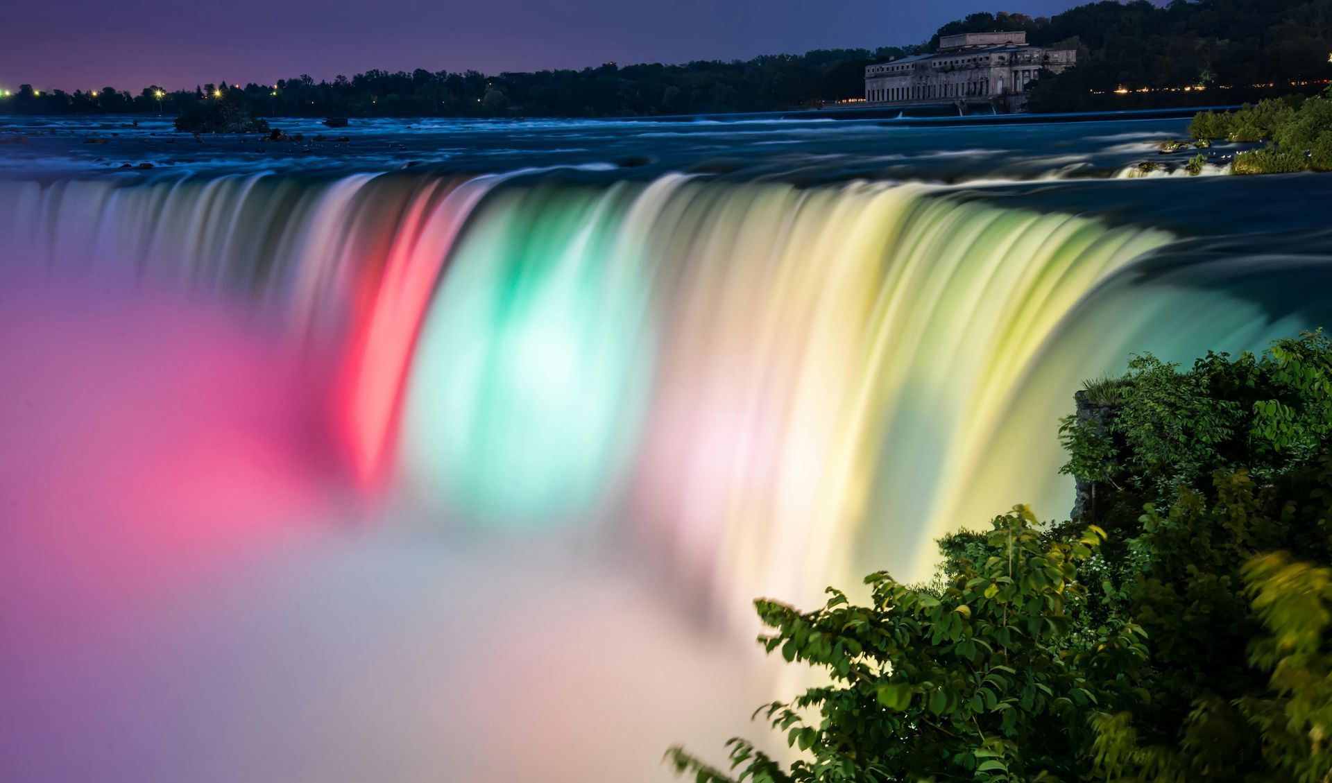 Niagara falls is lit up at night and looks like a rainbow.