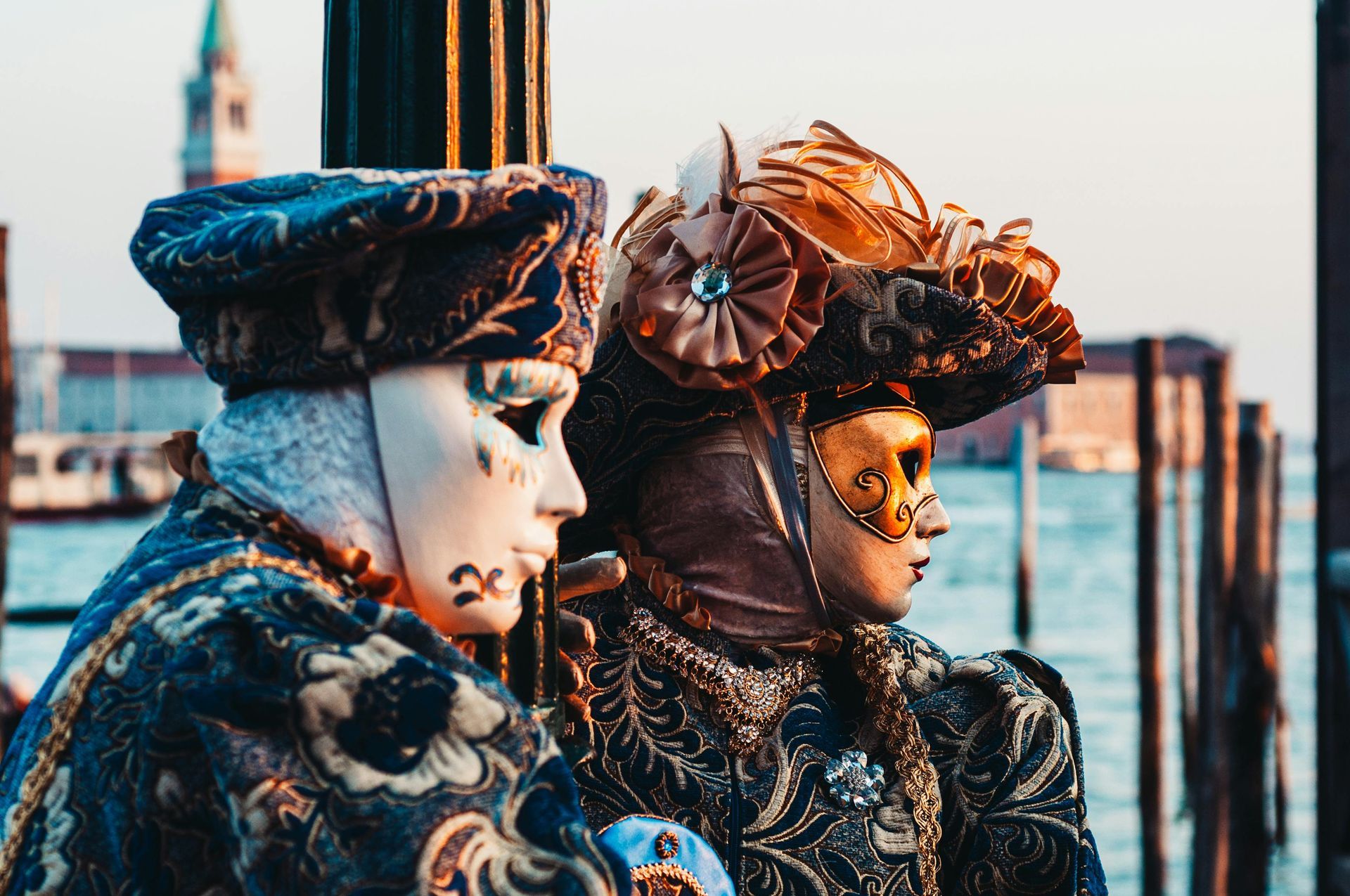 Two people wearing carnival masks are sitting next to each other on a pier.