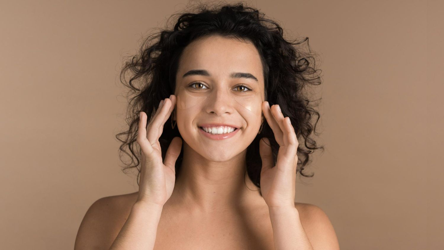 A woman with curly hair is smiling and touching her face.