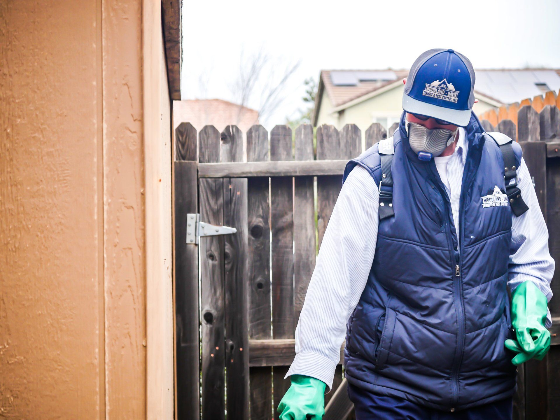 Worker in blue vest and cap cleaning near a wooden fence, wearing gloves and a face mask.