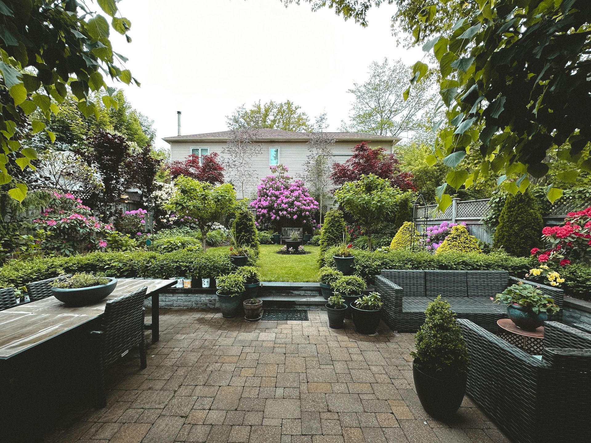 Lush garden patio with brick paving, benches, potted plants, and colorful flowering shrubs around a small building