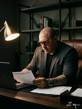 A person with a tattoo on their arm sits at a desk, reviewing a document with a laptop nearby under warm lamp lighting.