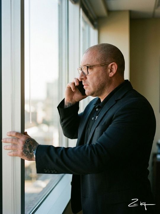A person wearing glasses and a dark blazer stands by a bright window, talking on a smartphone with a tattooed hand.