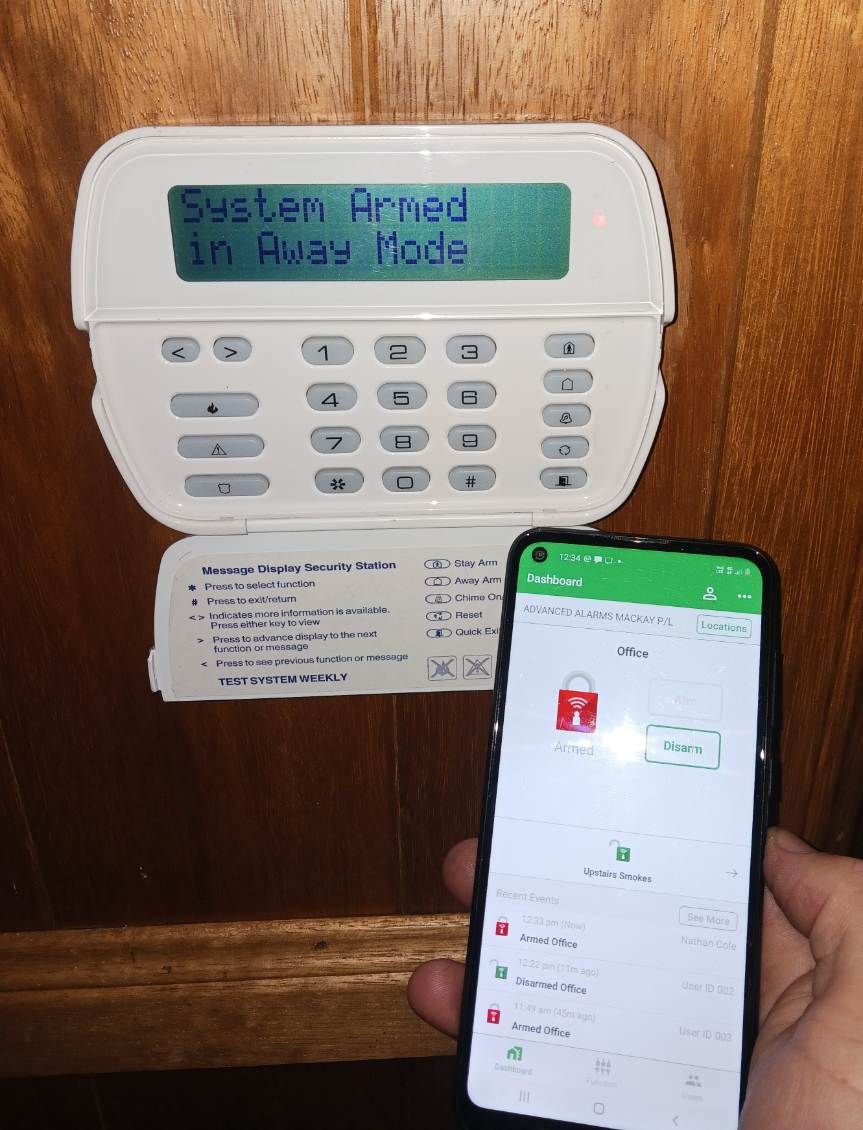 A Hand Holds A Smartphone Displaying A Security App Next To A Wall-mounted Alarm System Keypad. Both Show The System Is Armed In Away Mode — Advanced Alarms Mackay Pty Ltd In South Mackay, QLD