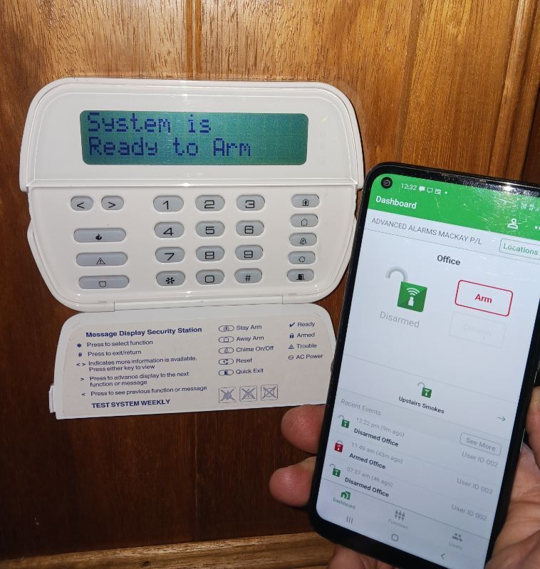 A Person Holding A Smartphone With A Security App Next To A Home Security System Keypad — Advanced Alarms Mackay Pty Ltd In South Mackay, QLD