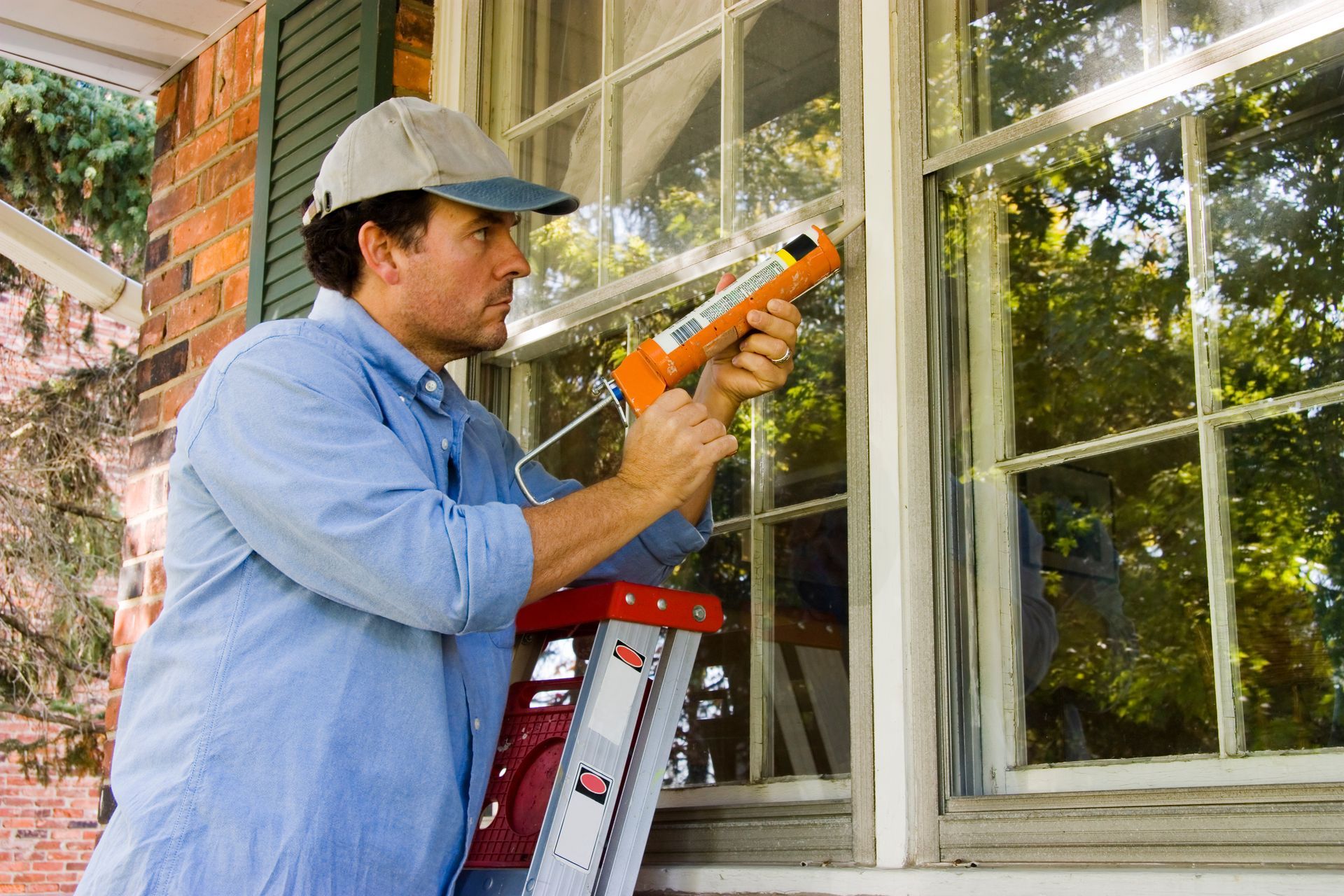 Man on a ladder caulking a window frame with a caulk gun.