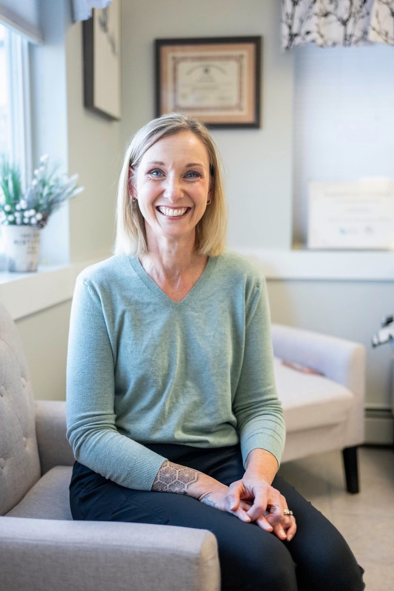 Woman sitting in a light grey chair, smiling, wearing a light blue sweater. Indoors.