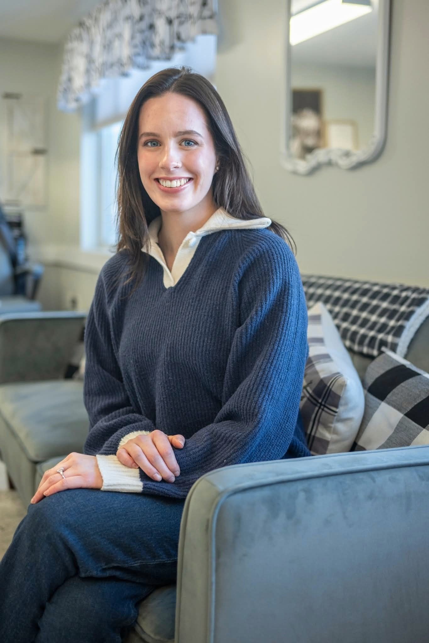 Woman smiling, seated on a gray couch in a living room, wearing a blue sweater and jeans.