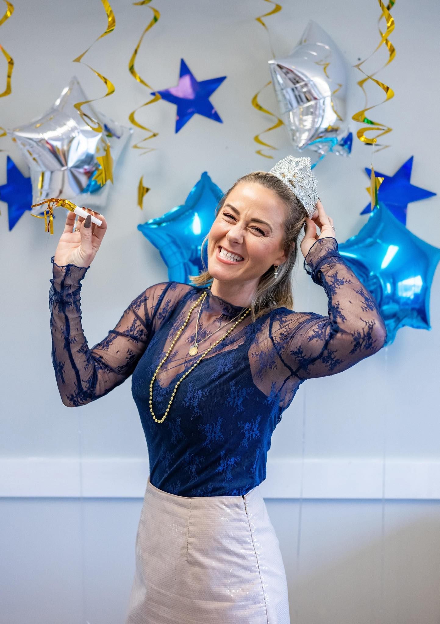 A woman wearing a tiara is standing in front of balloons and confetti.