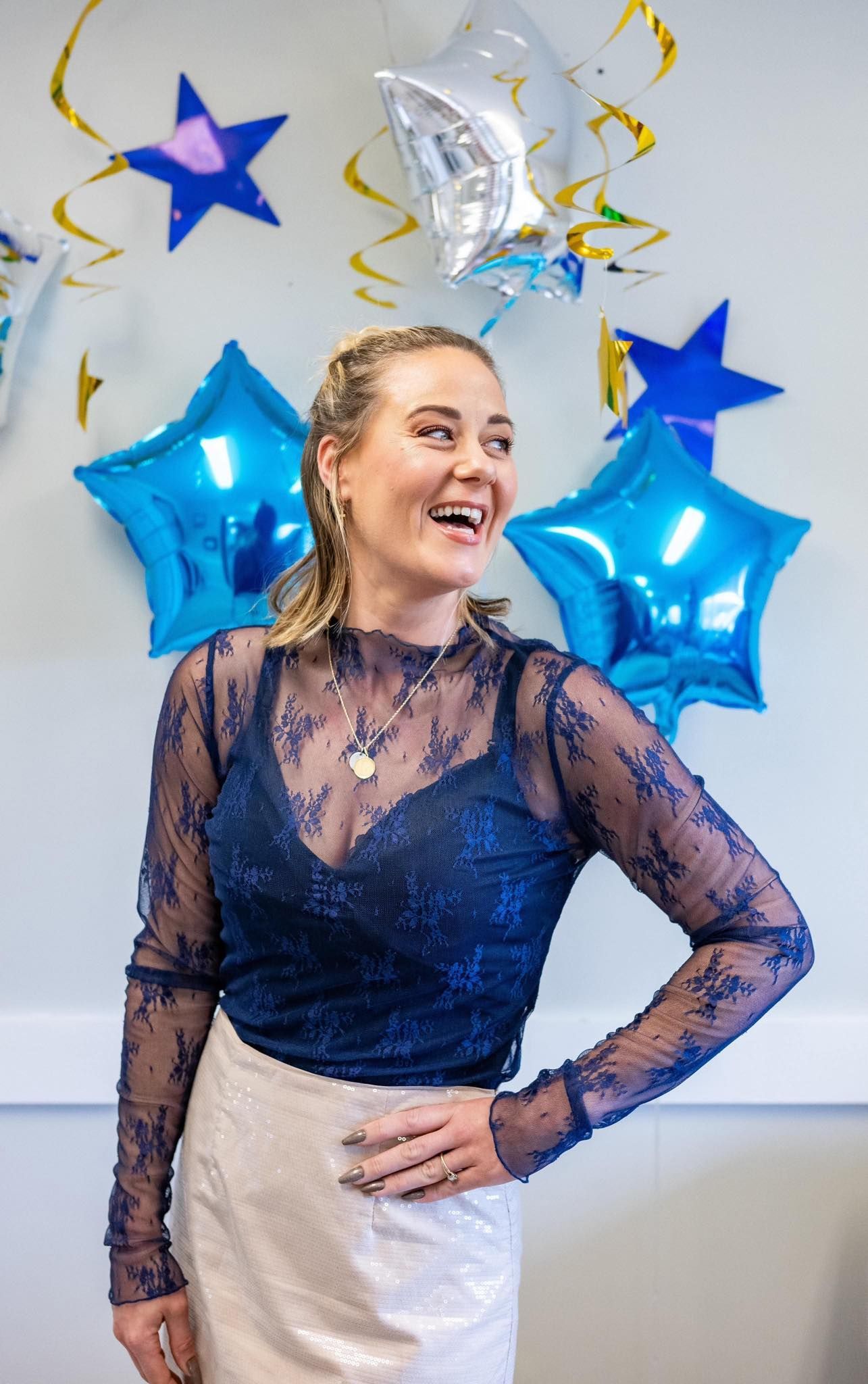 A woman in a blue top and white skirt is standing in front of blue balloons.