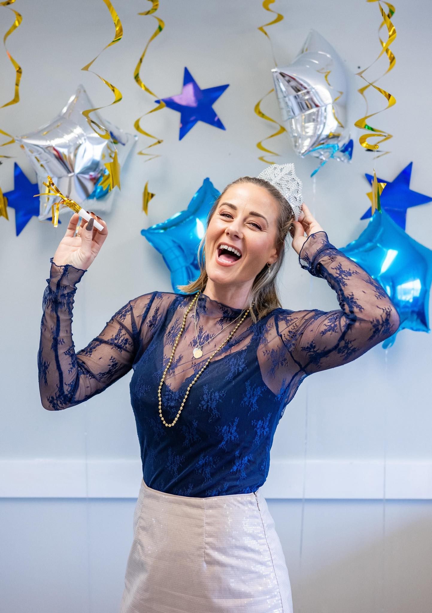 A woman wearing a tiara is standing in front of balloons and confetti.