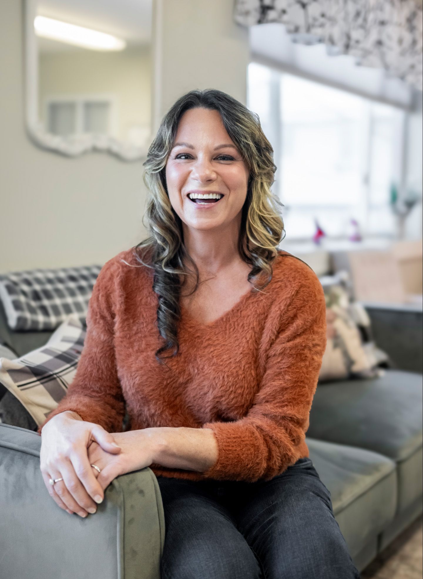 Woman smiling, seated on a gray couch; wearing an orange sweater and dark pants, indoors.