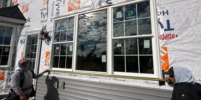 Two construction workers measuring a window on a house under construction. The white window has grids, and the house is covered in protective wrap.