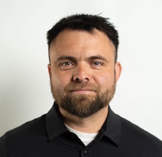 A man with a beard is wearing a black shirt and looking at the camera.