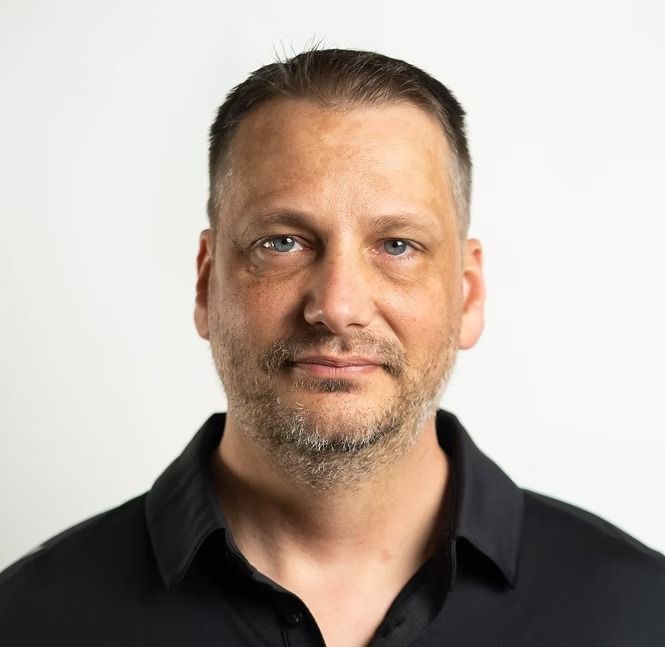 A man with a beard is wearing a black shirt and looking at the camera.