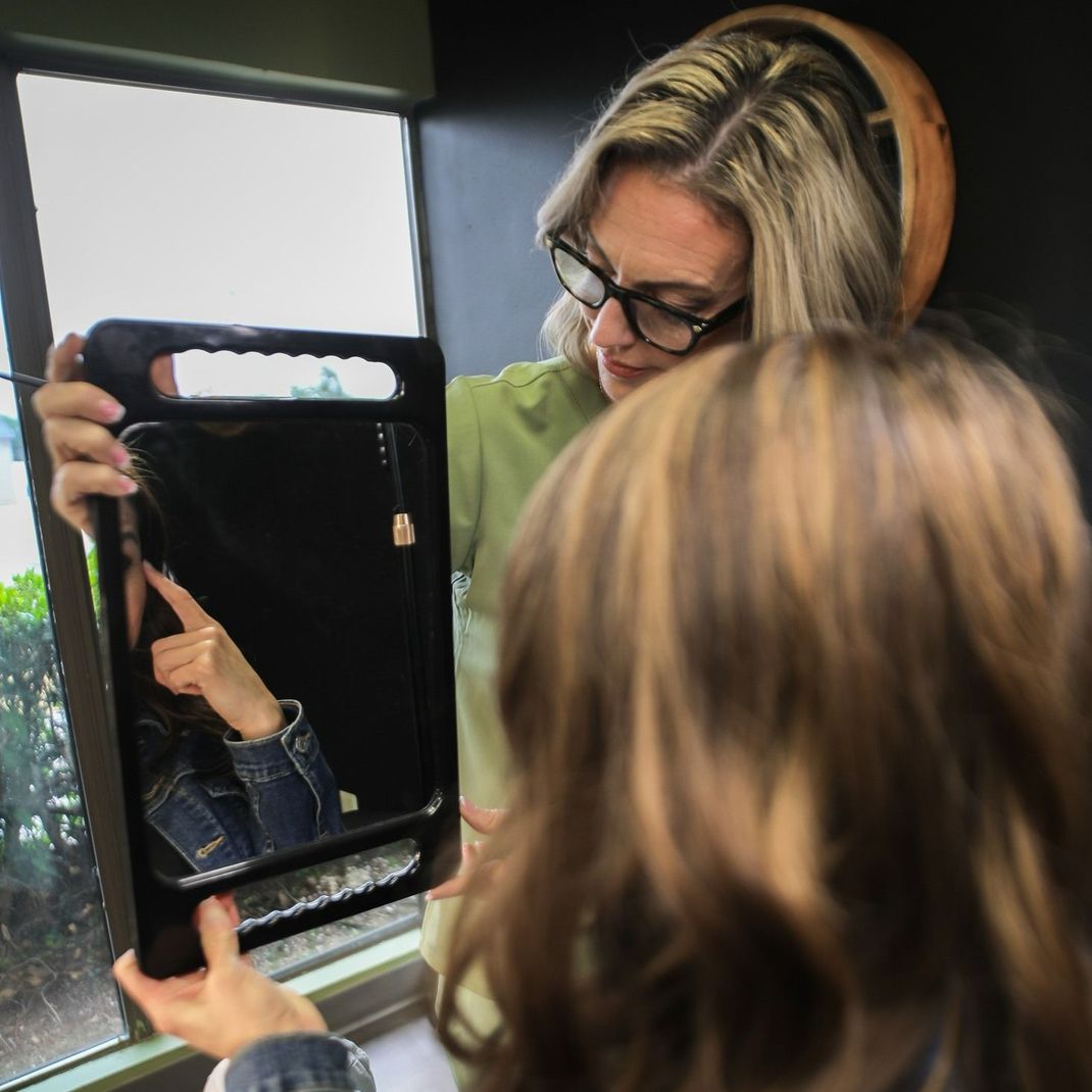 Woman holds up mirror, pointing to reflected image of another woman in denim jacket.