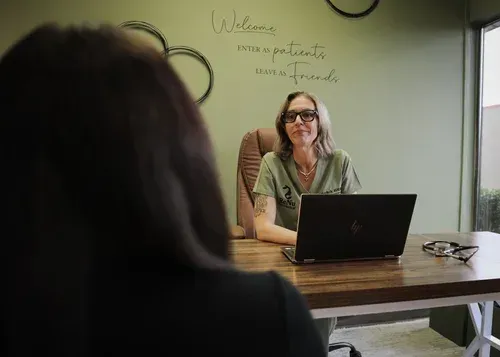 Doctor seated at desk, laptop open, speaking to a person out of focus. Green walls, text above.