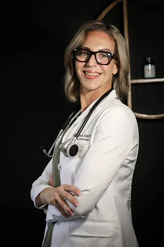 Woman in white lab coat with stethoscope, glasses. Smiles, arms crossed. Black background.