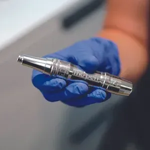 Person in blue glove holding a silver, pen-like medical device.