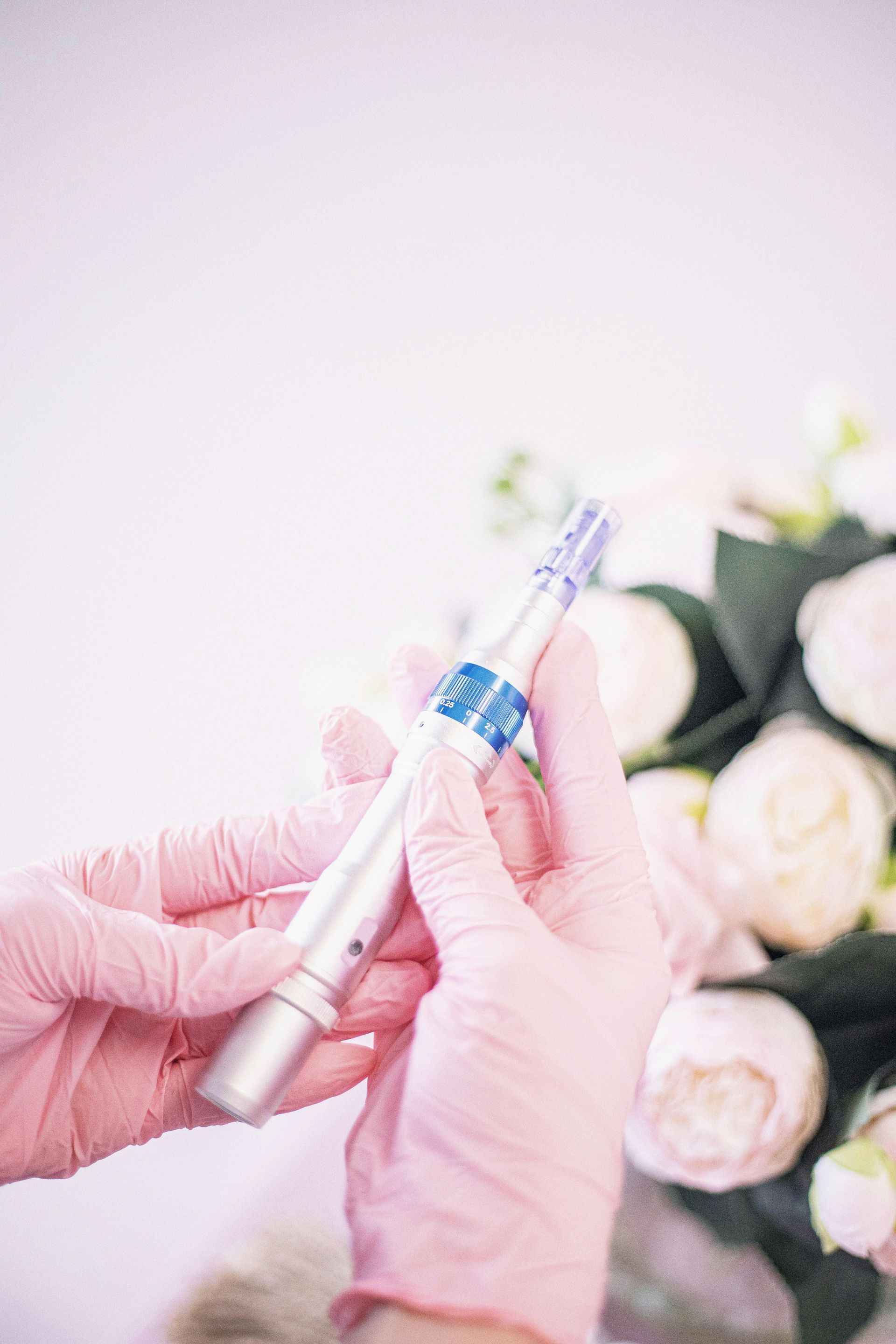 Hands in pink gloves holding a microneedling pen near white flowers.