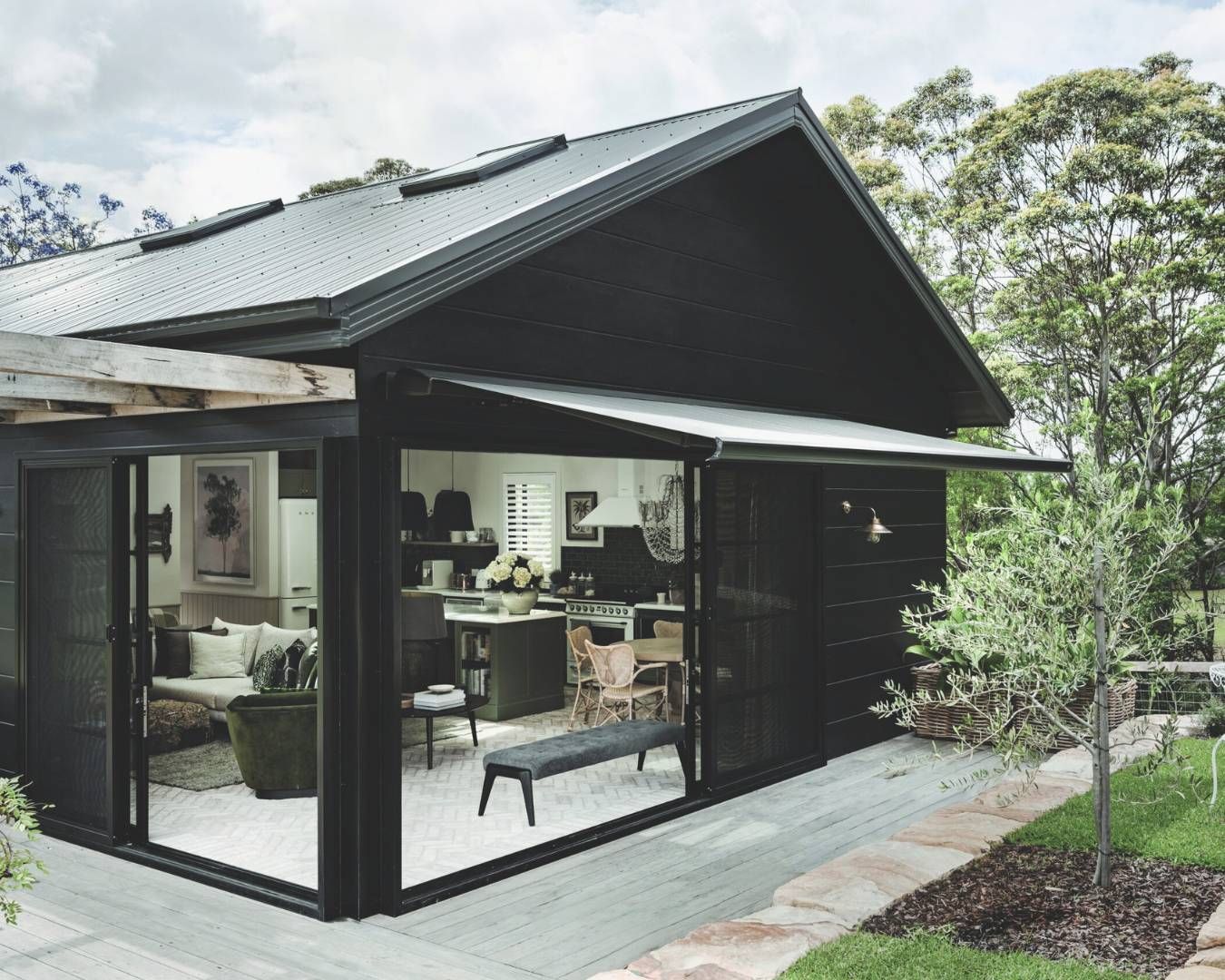 Spacious outdoor patio with a retractable awning, attached to a modern black house surrounded by greenery.