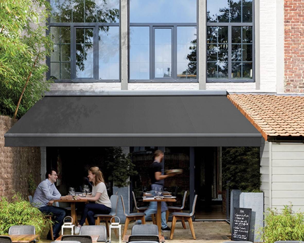 Outdoor dining area with a retractable awning providing shade, featuring a brick wall and greenery.