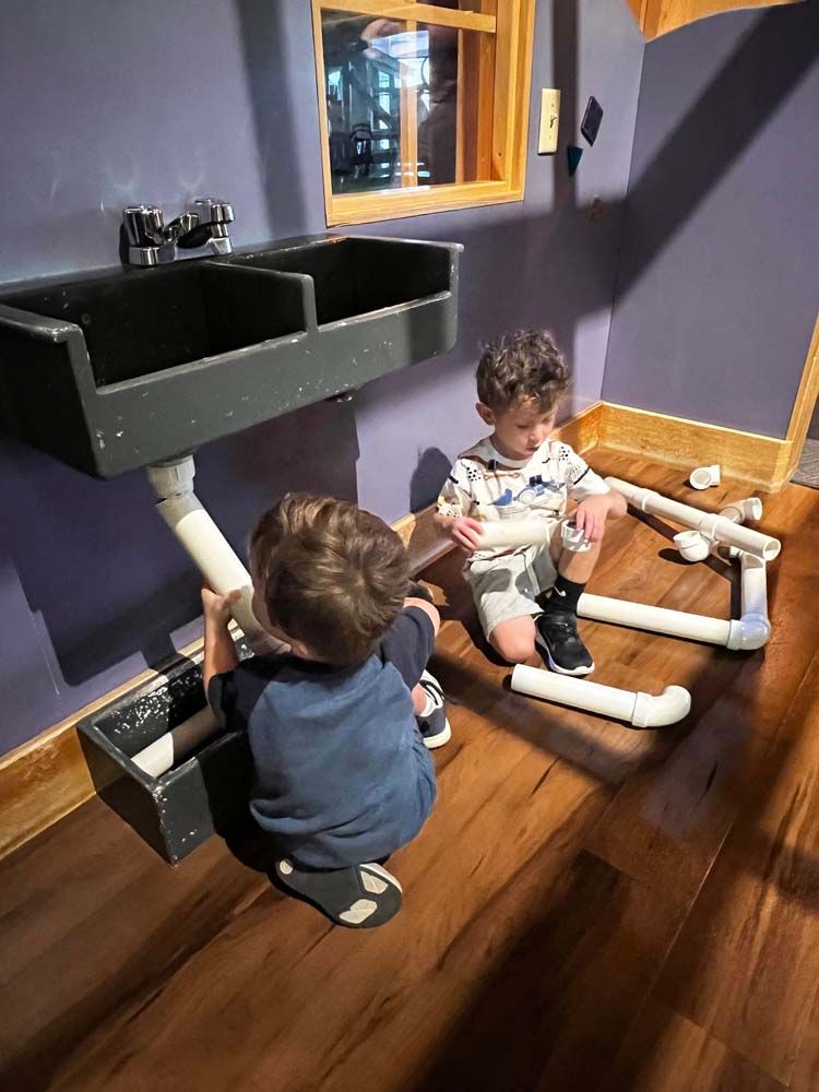 Two young boys are playing with pipes under a sink.